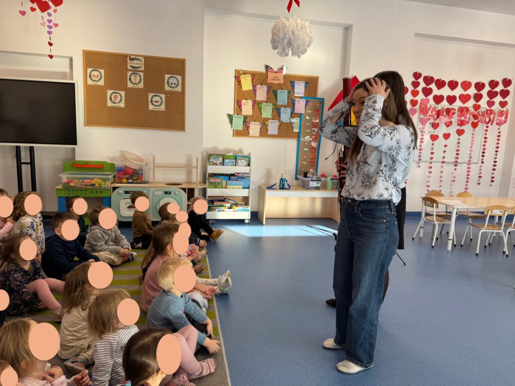 Pani trzyma się za głowę, opowiada do dzieci siedzących na dywanie