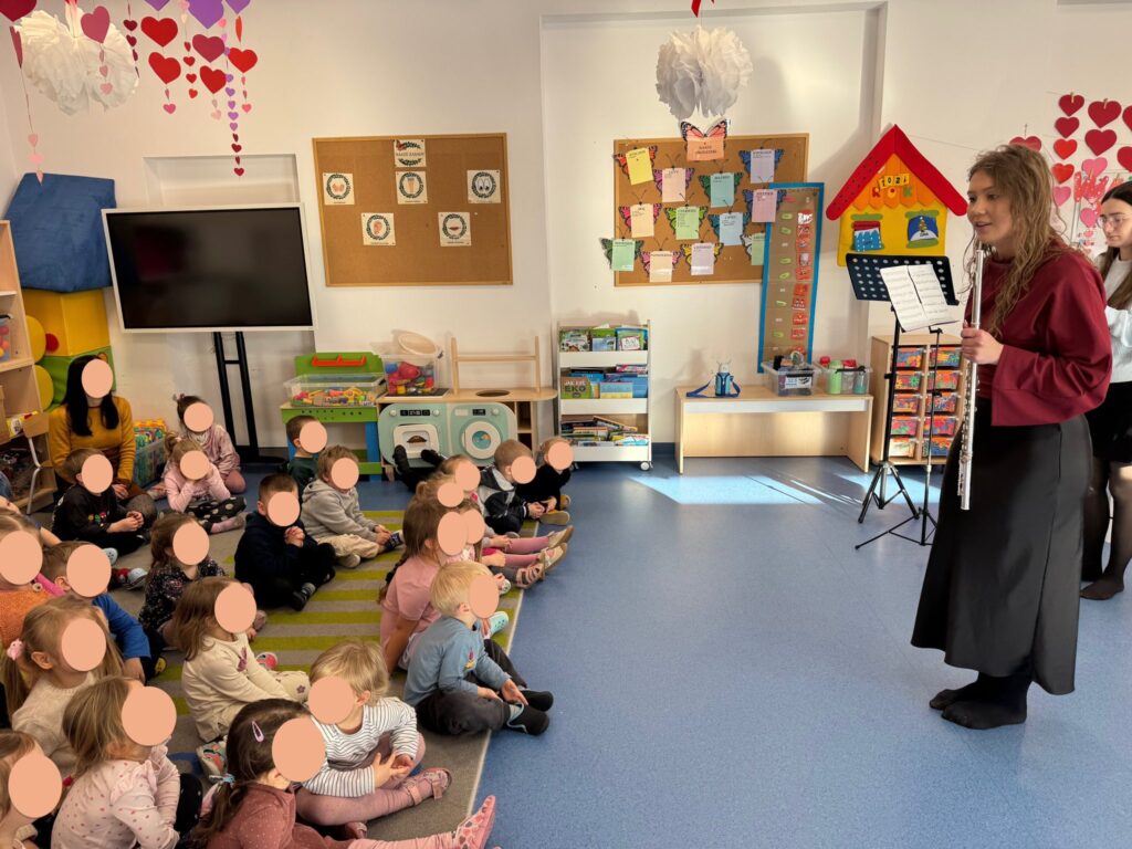 kobieta trzyma instrument muzyczny i mówi do dzieci siedzących na dywanie